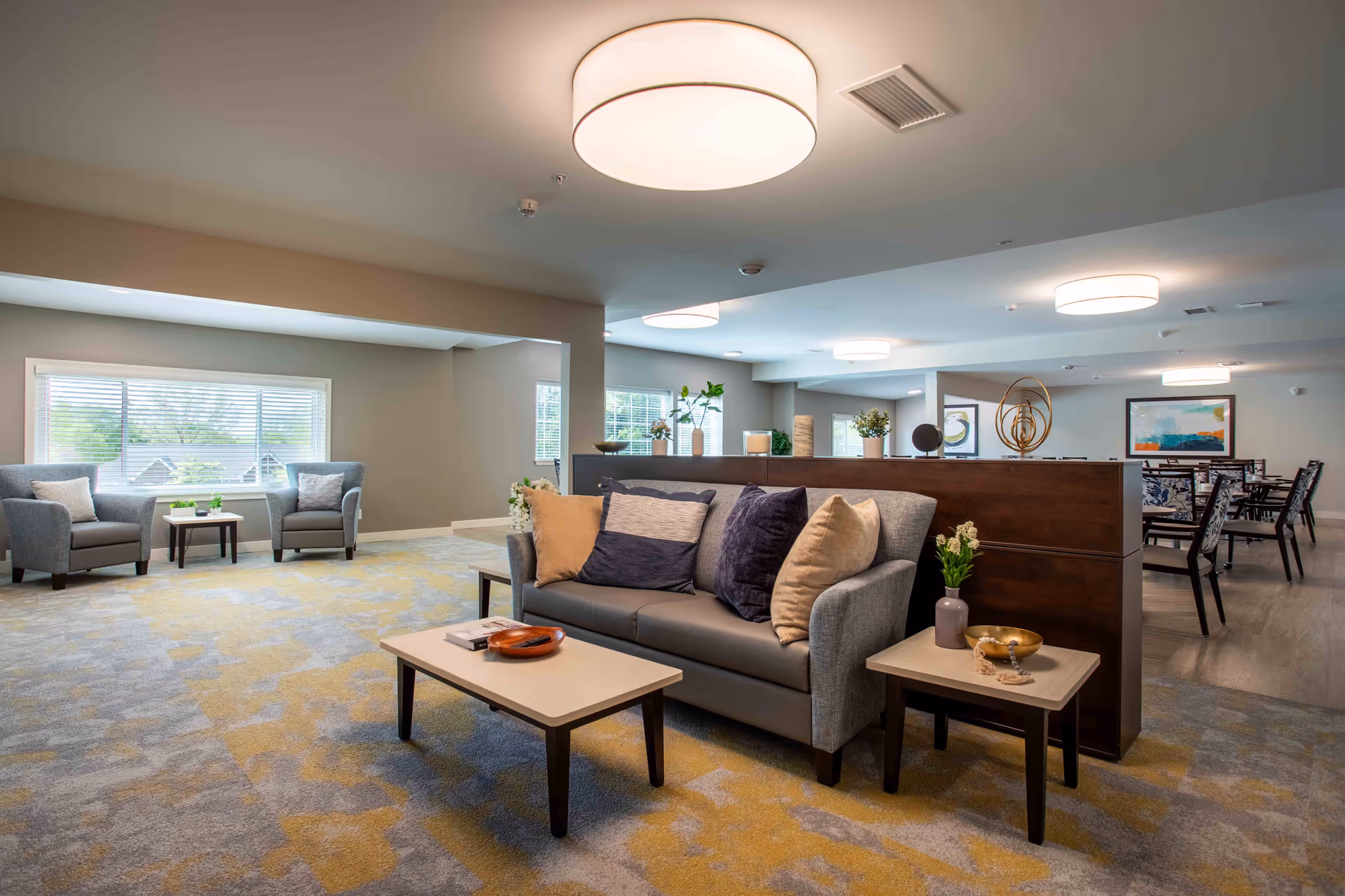 Common living area with gray sofa, armchairs, coffee tables, and a dining area beyond a partition.