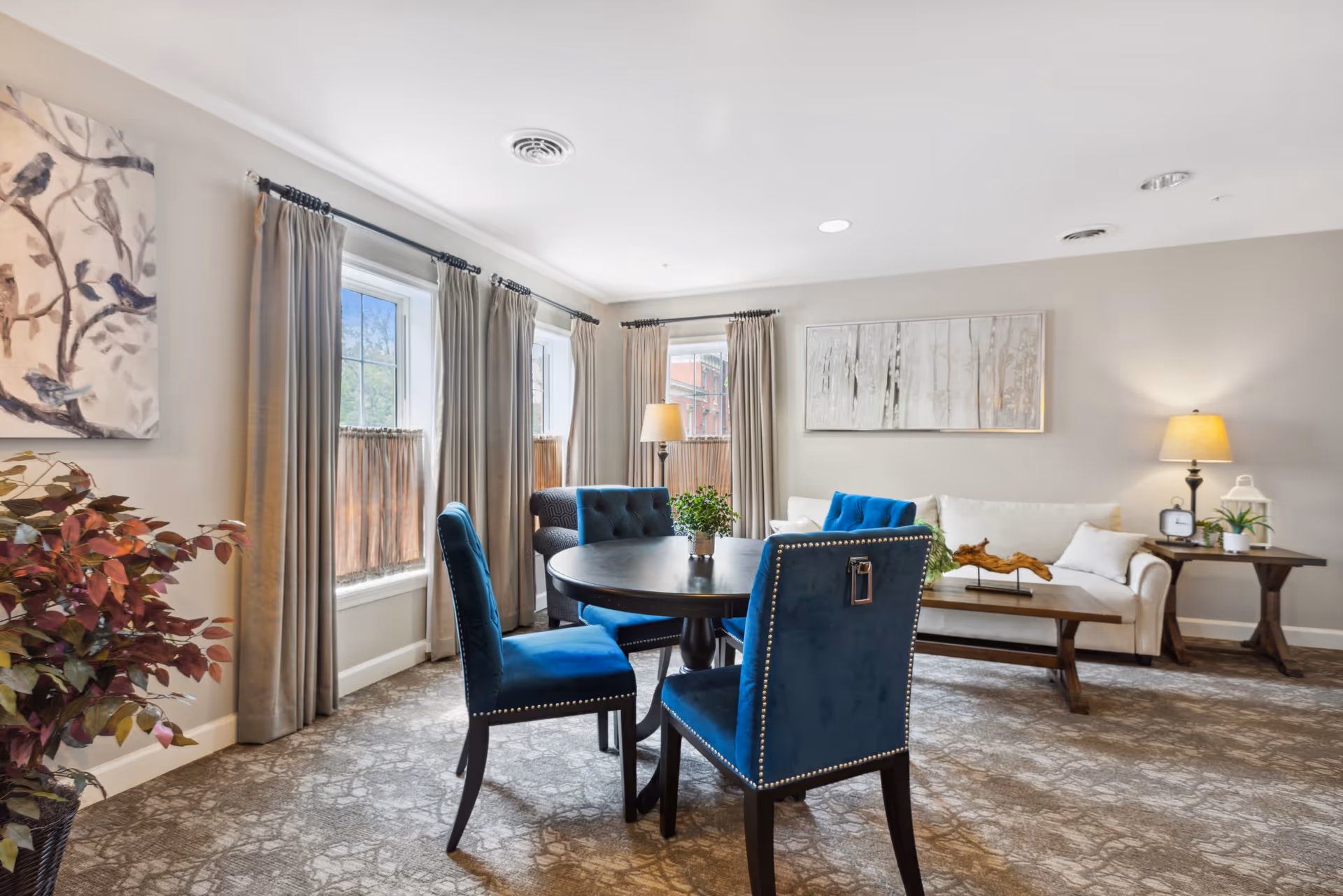 A bright and cozy living room in an assisted living facility featuring a round black table with four blue upholstered chairs, a white sofa with blue and white cushions, a wooden coffee table, two side tables with lamps and plants, large windows with beige curtains, and decorative wall art including a painting of birds and a forest scene.