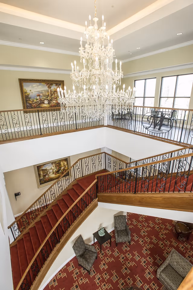 View from above of an elegant two-story interior space with a large crystal chandelier hanging from the ceiling. The area features a red carpeted staircase with ornate black wrought iron railings, patterned red carpet flooring, two gray upholstered chairs with a small table between them, and large windows letting in natural light. There are framed paintings on the walls and a decorative metal 'M' on the railing.
