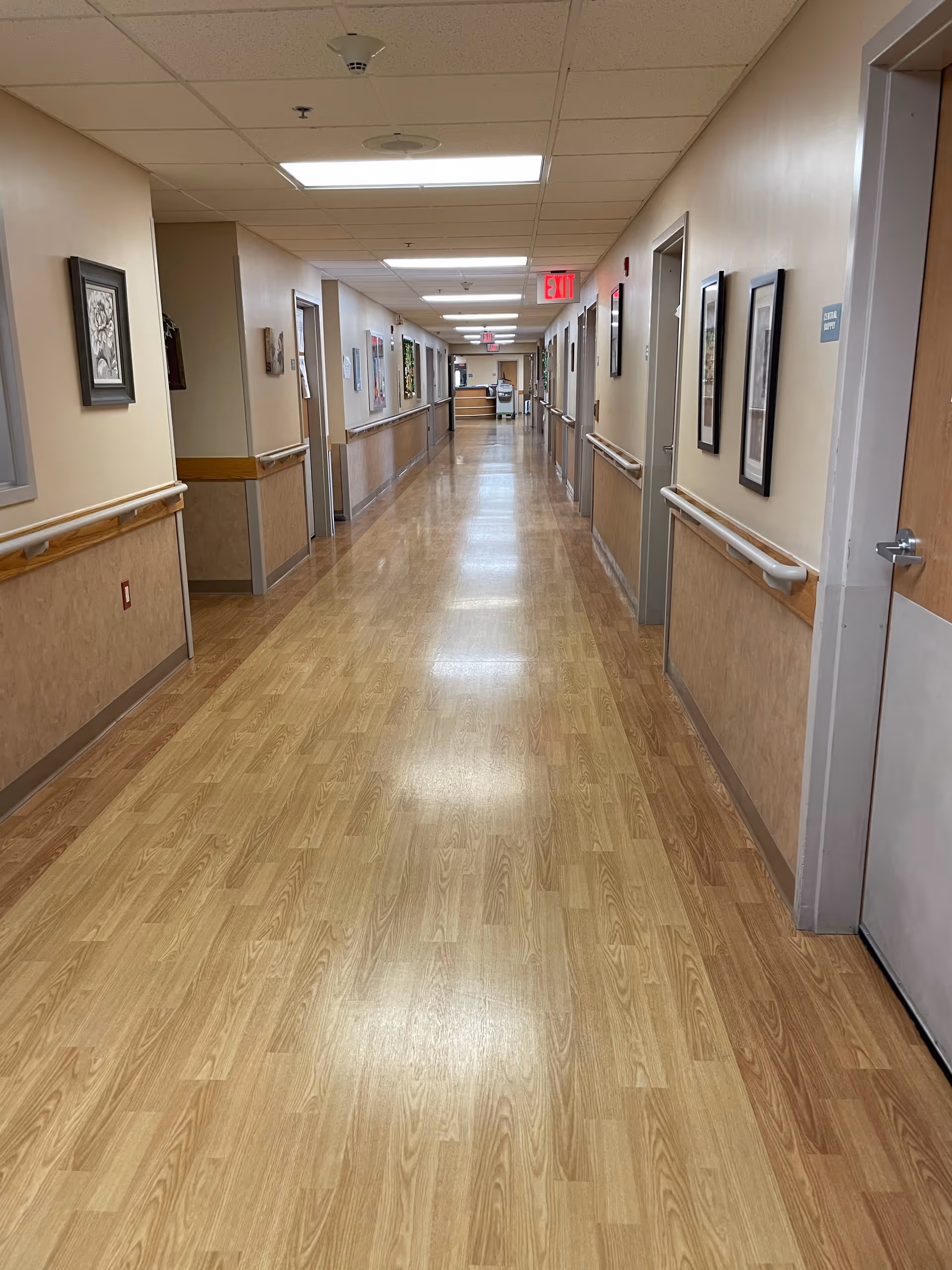 Long, well-lit interior hallway in a senior living facility with handrails, framed artwork, doors, and an exit sign.
