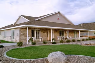 Single-story assisted living facility building with a covered porch featuring several white chairs. The building has a brick exterior with beige siding and a well-maintained lawn with landscaping including small bushes and a large rock in the foreground.