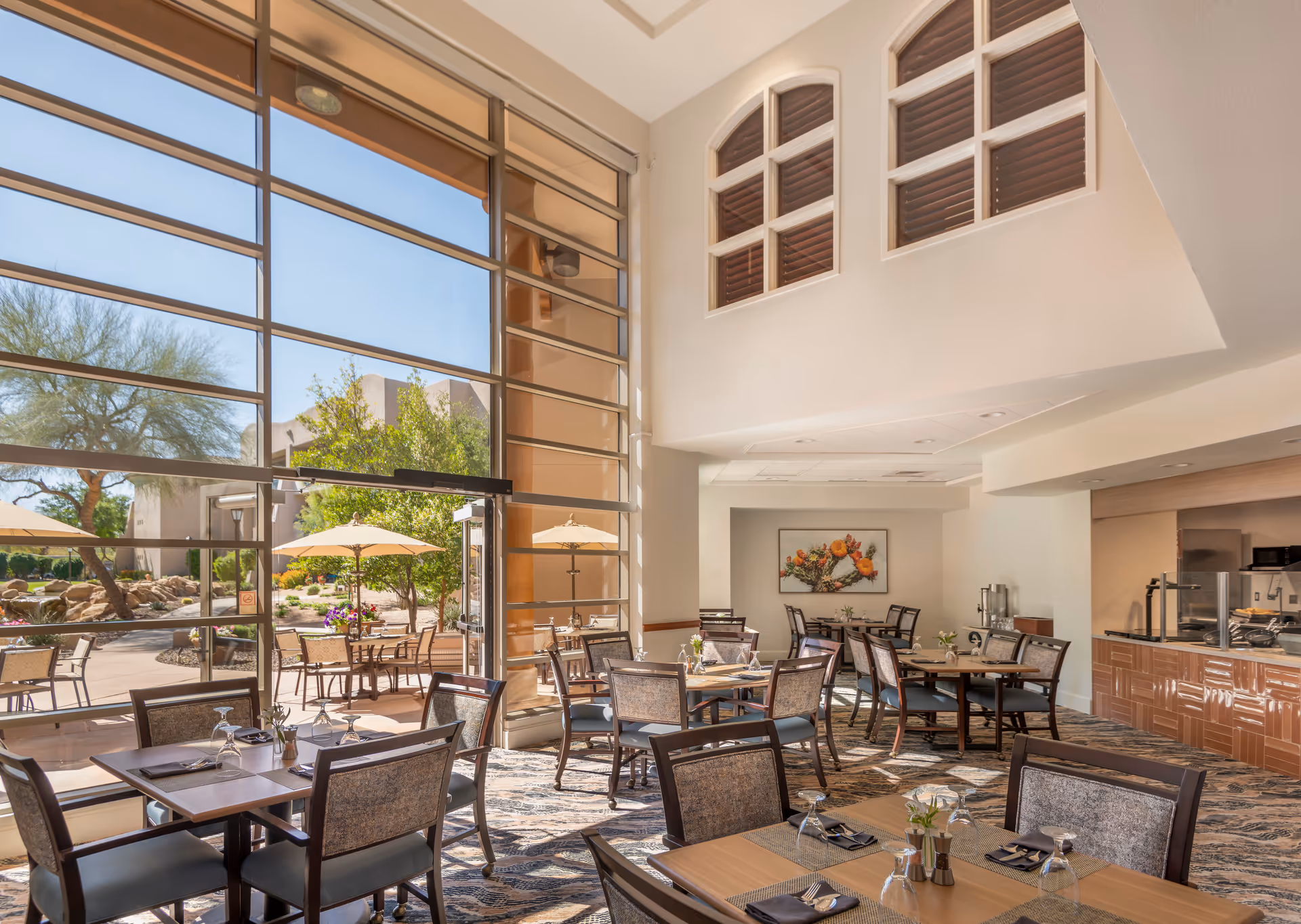 Bright dining area with multiple tables and chairs, set with glasses and napkins. Large floor-to-ceiling windows provide a view of an outdoor patio with tables, chairs, and umbrellas. The interior features light-colored walls, a patterned carpet, and a serving counter on the right side. A framed floral artwork hangs on the far wall.