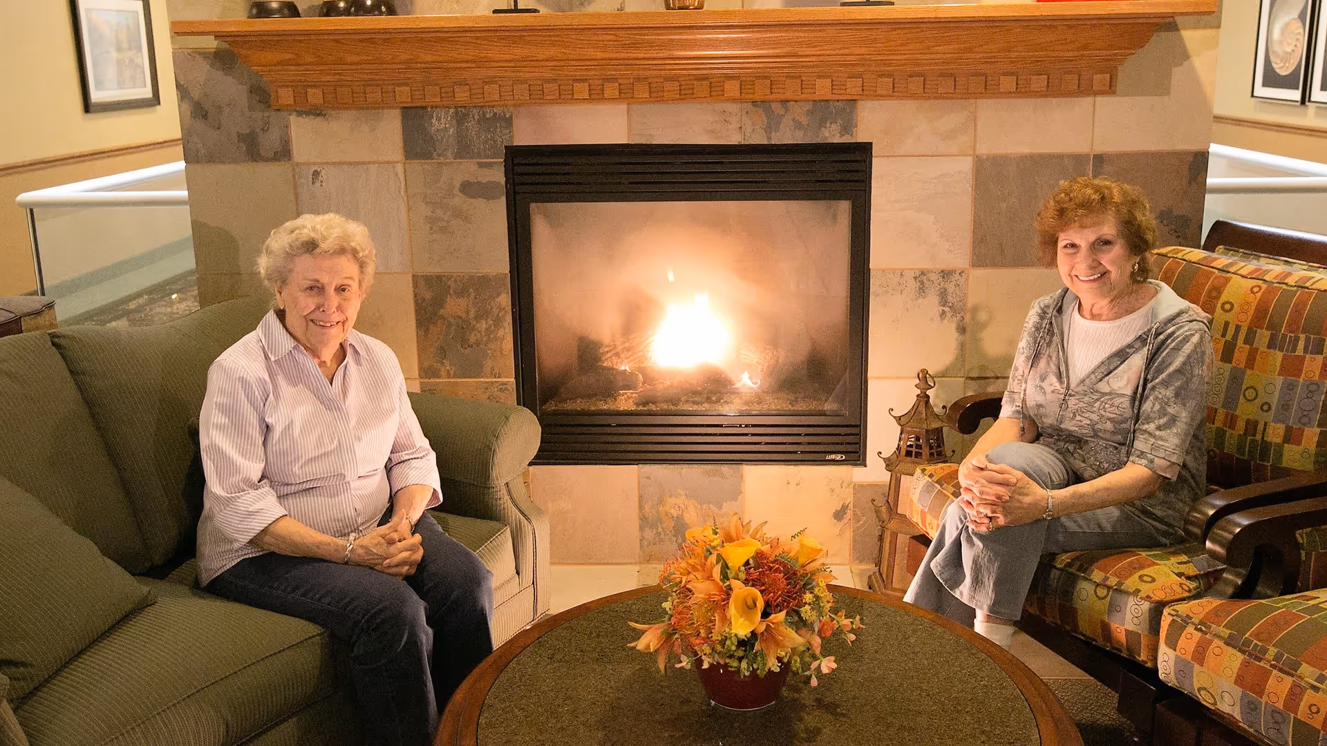 Two elderly women sitting comfortably in a cozy living room area with a lit fireplace behind them. One woman is seated on a green couch and the other on a patterned armchair. A round table with a floral arrangement is in front of them.
