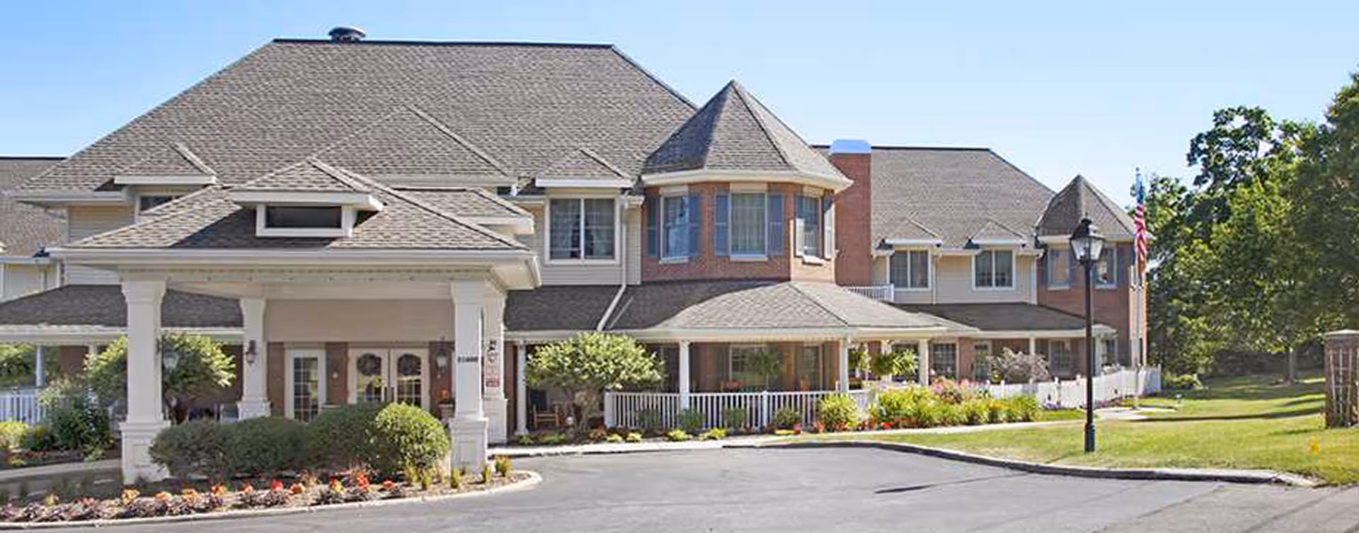 Exterior view of Bickford of Rocky River, a large senior living facility with a covered entrance, multiple windows, and a well-maintained garden and lawn area under a clear blue sky.