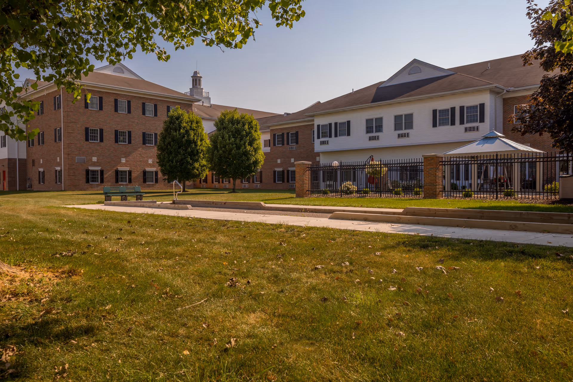 Exterior view of Shepherd of the Valley - Howland facility showing a large brick and white building with multiple windows, a fenced patio area, green grass lawn, trees, and a clear sky.