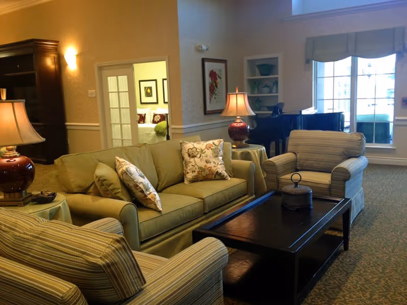 A cozy living room with a green sofa and two striped armchairs arranged around a dark wooden coffee table. There are two table lamps with red bases on side tables, a framed floral picture on the wall, a large window with a valance, and a black grand piano in the corner. A doorway leads to a bedroom with a bed and framed pictures visible.