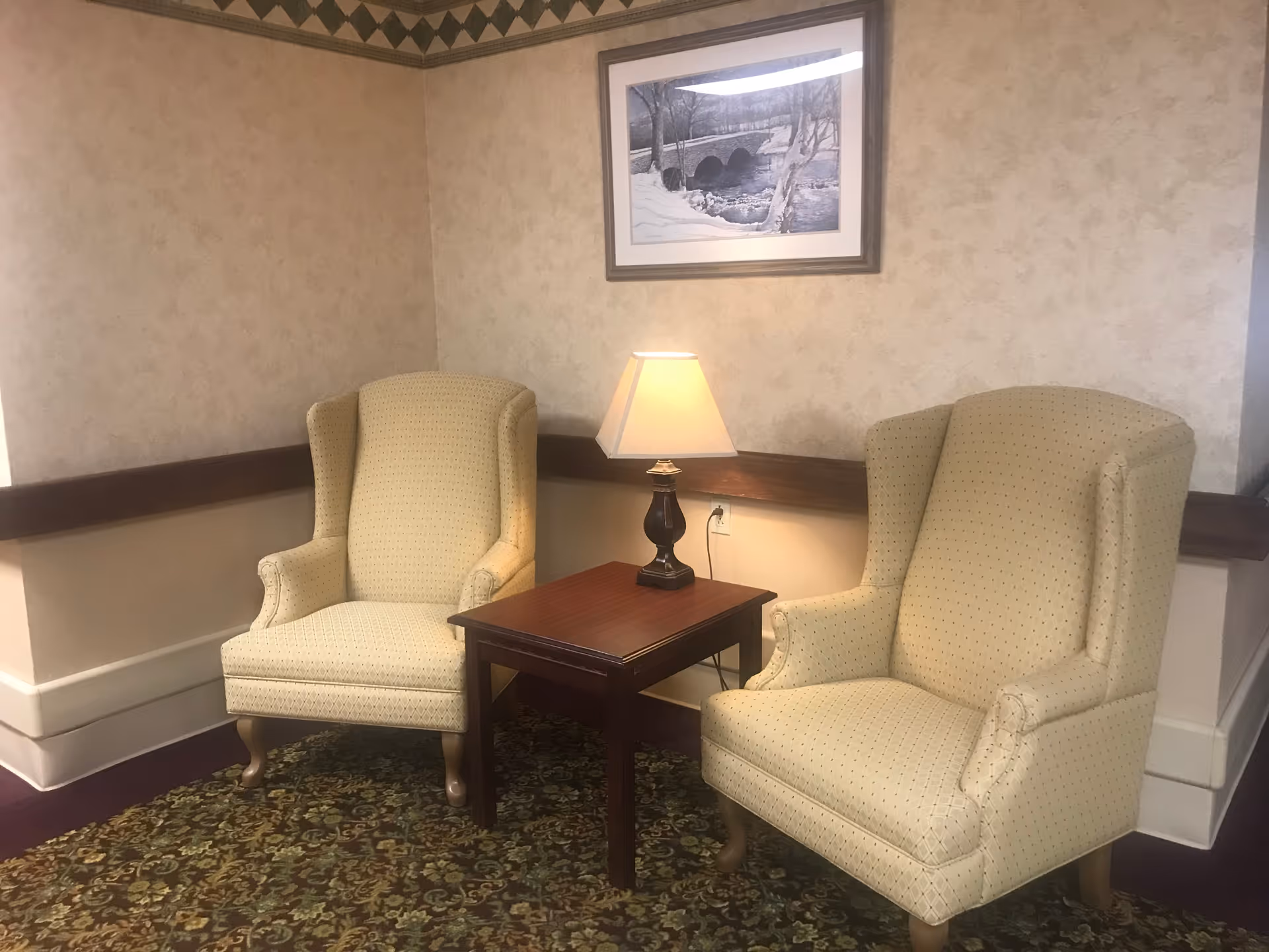 Two beige upholstered armchairs with wooden legs are positioned on either side of a small wooden table with a lamp on it. The setting is indoors with patterned carpet and wallpapered walls. A framed black and white picture of a bridge over a stream hangs on the wall above the chairs.