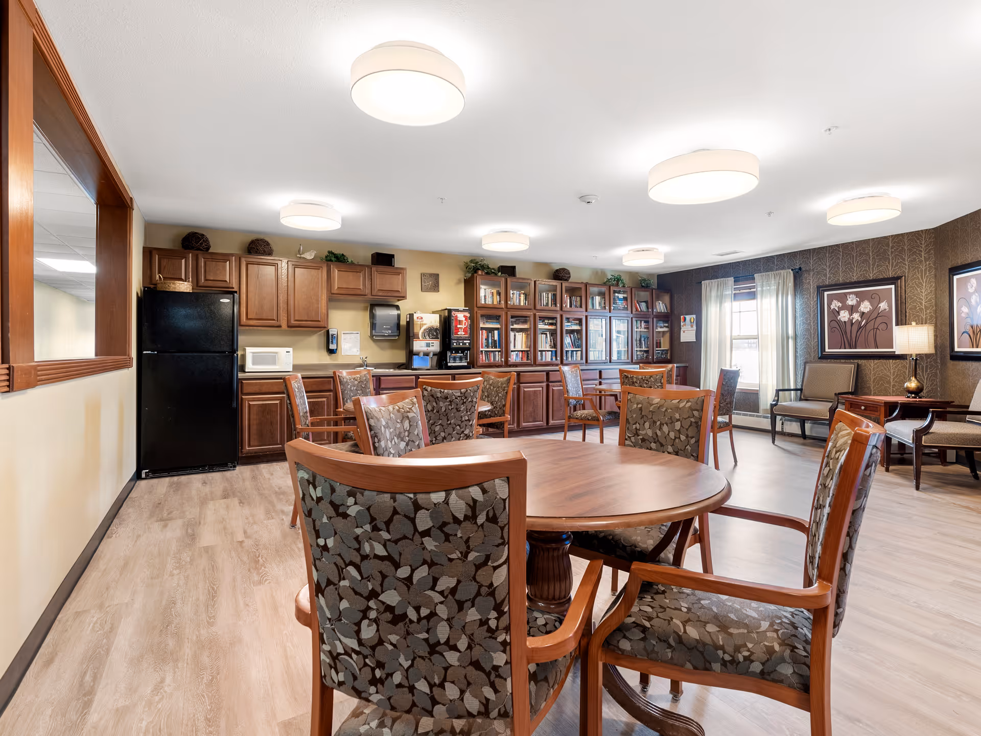 A well-lit common area in a senior living facility featuring round wooden tables with patterned cushioned chairs, a kitchenette with a black refrigerator, microwave, coffee machine, and wooden cabinets. There is a large bookshelf filled with books along the back wall, windows with light curtains, and framed floral artwork on the walls. The room has light wood flooring and modern ceiling lights.