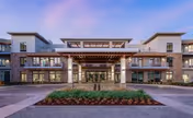 Front exterior view of a modern senior living facility building with large windows, a covered entrance supported by columns, and landscaped greenery in front under a clear sky at dusk.