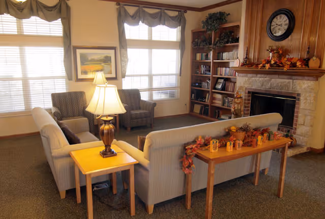 A cozy living room with two beige sofas arranged around a wooden side table with a lamp. There are two armchairs near large windows with blinds and valances. A wooden bookshelf filled with books and decorative items stands next to a stone fireplace with a clock and autumn-themed decorations on the mantel.