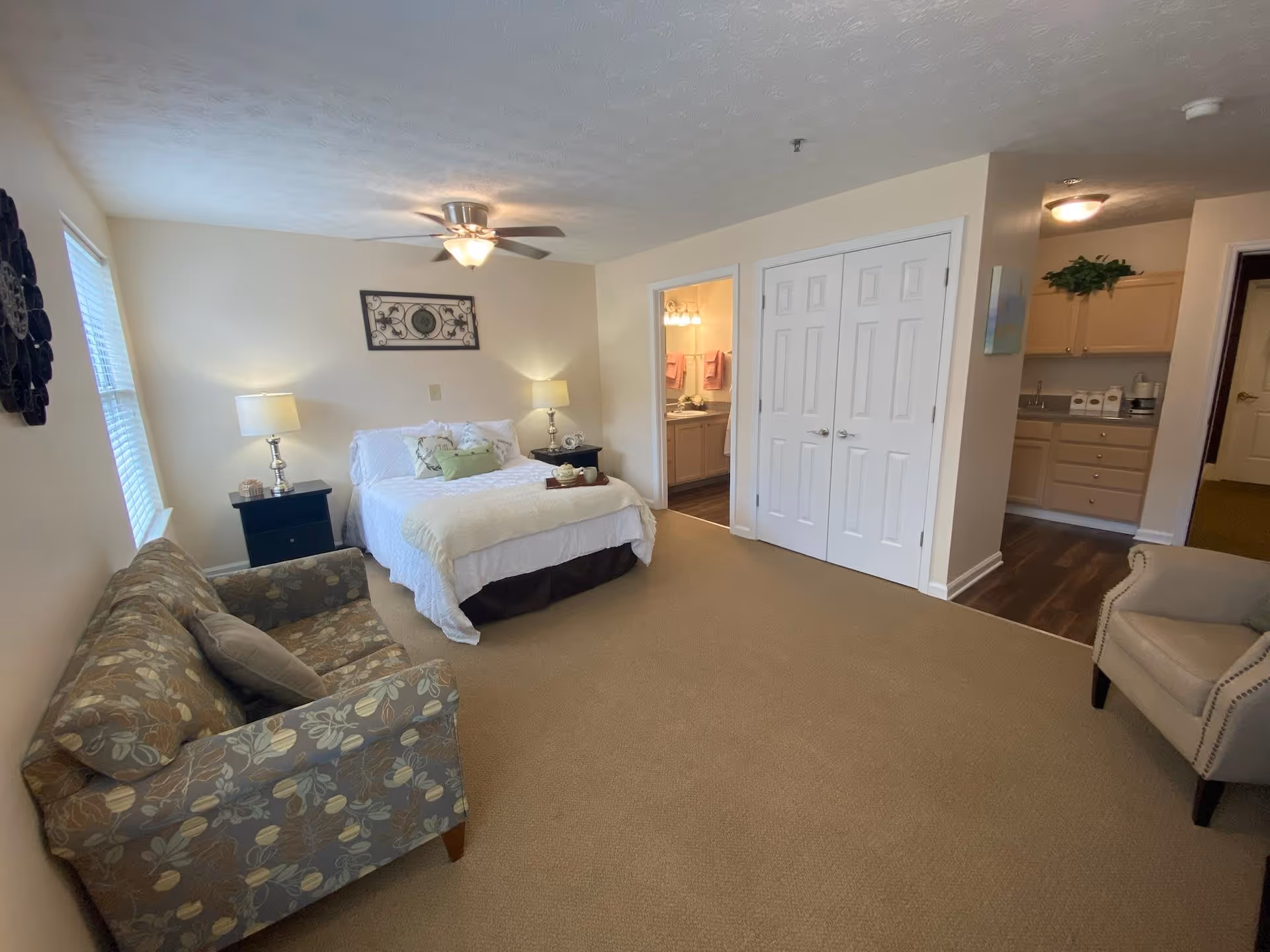 A spacious bedroom with a bed centered against the back wall, flanked by two nightstands with lamps. To the left is a floral-patterned couch, and to the right is a beige armchair. There is a ceiling fan with a light fixture above the bed. A doorway on the right leads to a bathroom, and another open area reveals a kitchen with cabinets and a countertop.
