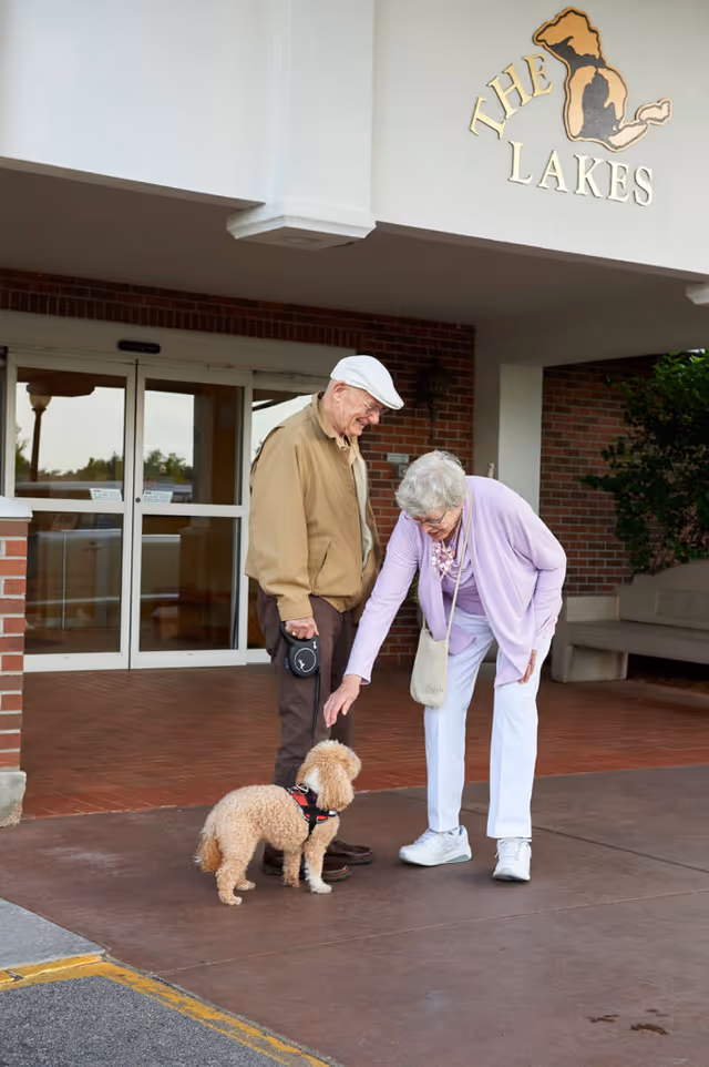 An elderly man and woman standing outside the entrance of a building named 'The Lakes.' The man is holding a small dog on a leash, and the woman is bending down to pet the dog. The building has brick walls and glass doors.