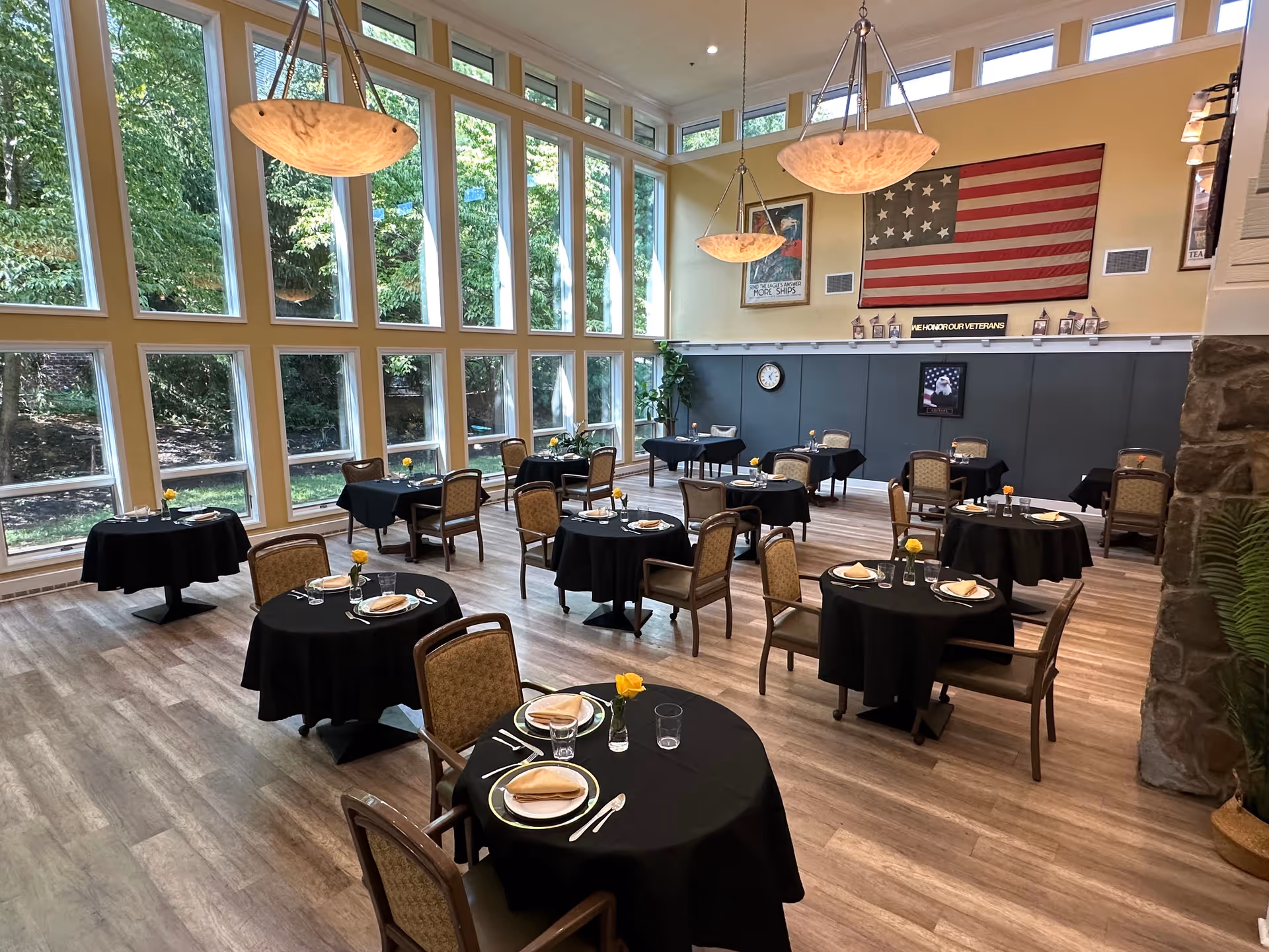 A dining room with multiple round tables covered with black tablecloths, each set with plates, napkins, glasses, and yellow flowers in small vases. The room features large floor-to-ceiling windows letting in natural light, wooden flooring, and three hanging pendant lights. An American flag and patriotic posters are displayed on the far wall.