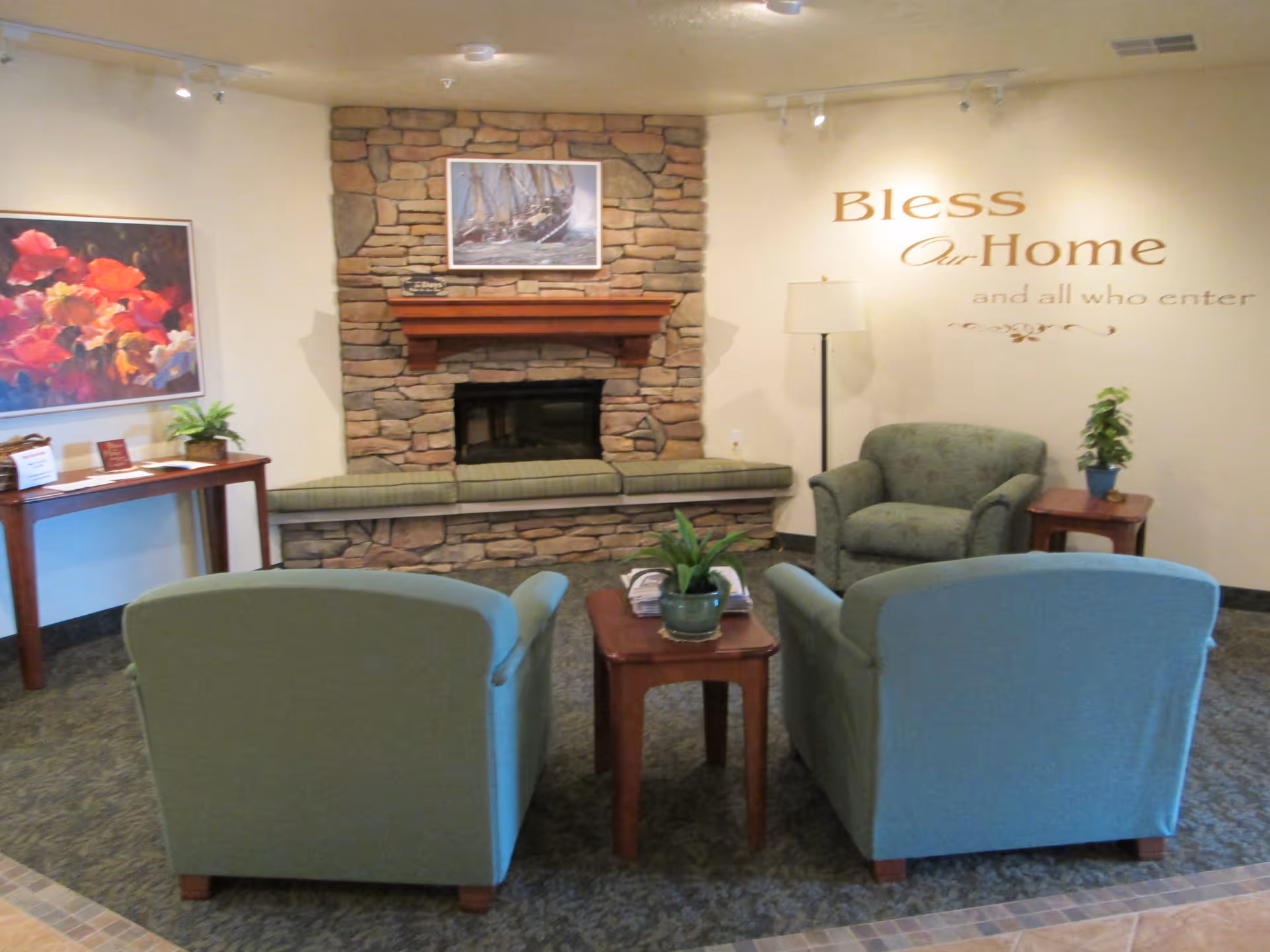 A cozy lounge with four upholstered armchairs around a small table facing a stone fireplace beneath a wall decal that reads "Bless Our Home".