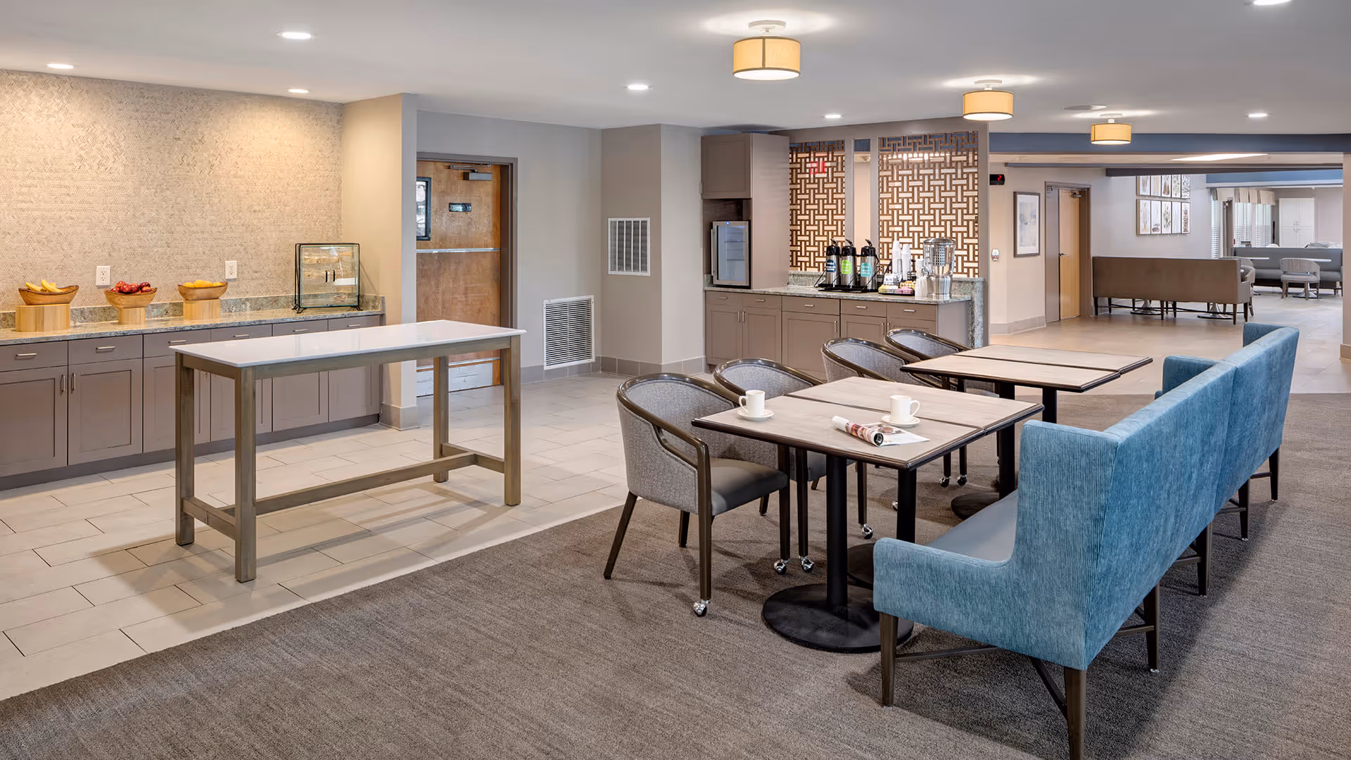 A spacious and well-lit common area in a senior living facility featuring a seating area with tables, chairs, and a blue upholstered bench. There is a counter with coffee and beverage dispensers, and a small table in the center of the room. The walls are light-colored, and the floor is a combination of tile and carpet. The area appears clean and inviting.