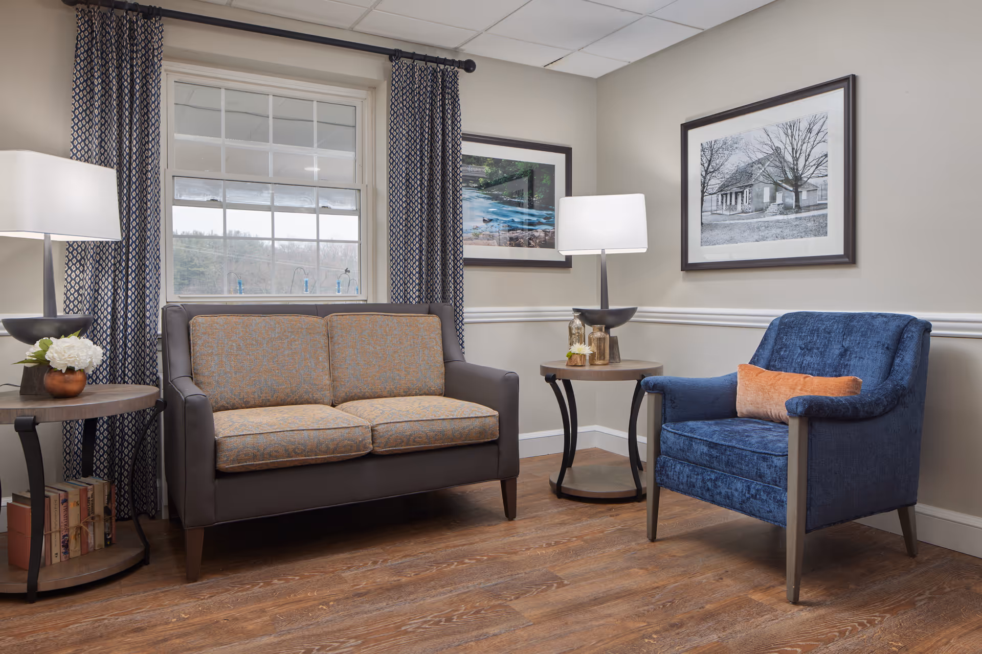 A cozy living room area with a brown loveseat featuring patterned cushions, a blue armchair with an orange pillow, two round wooden side tables each with a lamp, and framed artwork on the beige walls. A window with patterned curtains lets in natural light, and the floor is wooden.