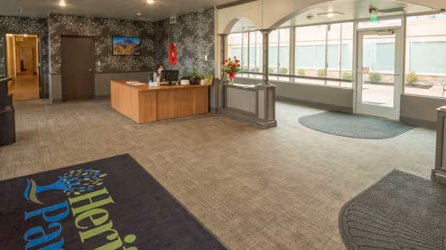 Reception area of Heritage Park Healthcare and Rehabilitation featuring a wooden front desk with a computer and a staff member seated behind it. The room has carpeted floors with mats near the entrance, large windows allowing natural light, and a hallway leading to other parts of the facility.