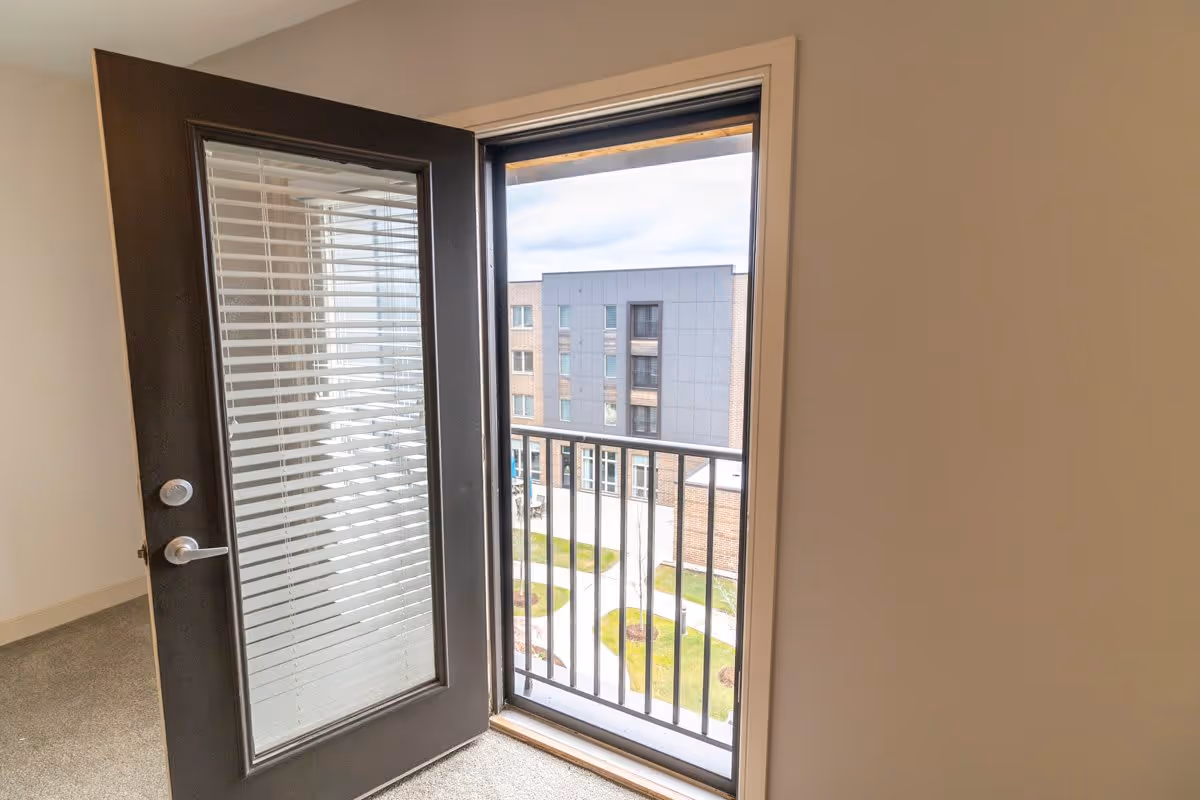 Open glass door with built-in blinds leading to a small balcony with a black metal railing, overlooking a courtyard and a modern multi-story building.