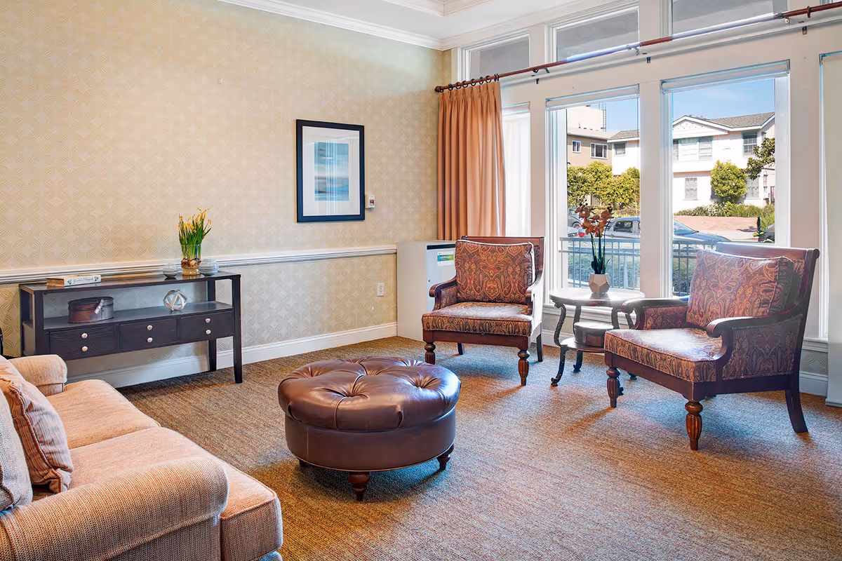 A cozy living room area with a beige sofa on the left, two patterned armchairs on the right, and a round brown leather ottoman in the center. There is a small dark wooden side table with drawers against the wall, decorated with a vase of flowers and other small items. Large windows with white frames and peach-colored curtains let in natural light and show a view of neighboring buildings and greenery outside.