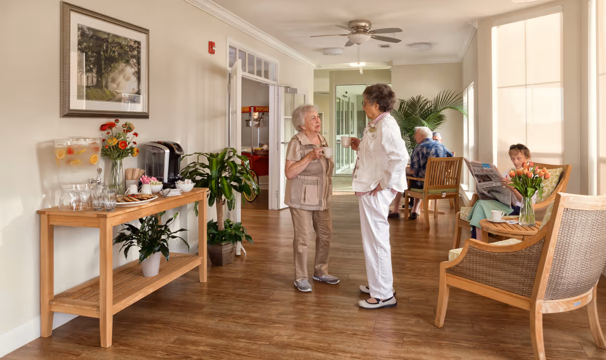 A bright common area in a senior living facility with wooden flooring and light-colored walls. Two elderly women are standing and chatting while holding cups. In the background, two other elderly individuals are seated on wooden chairs, one reading a newspaper. A wooden table on the left side holds a water dispenser with lemon slices, a coffee maker, cups, and some snacks. There are plants and flowers placed around the room, and large windows with blinds allow natural light to fill the space.