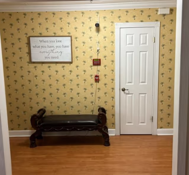 A small interior area with a wooden bench with dark leather upholstery placed against a wall covered in yellow wallpaper with a small palm tree pattern. A white door with a silver handle is on the right side of the wall. Above the bench is a framed sign that reads, 'When you love what you have, you have everything you need.' There are also a fire alarm pull station and a fire alarm bell mounted on the wall between the bench and the door.