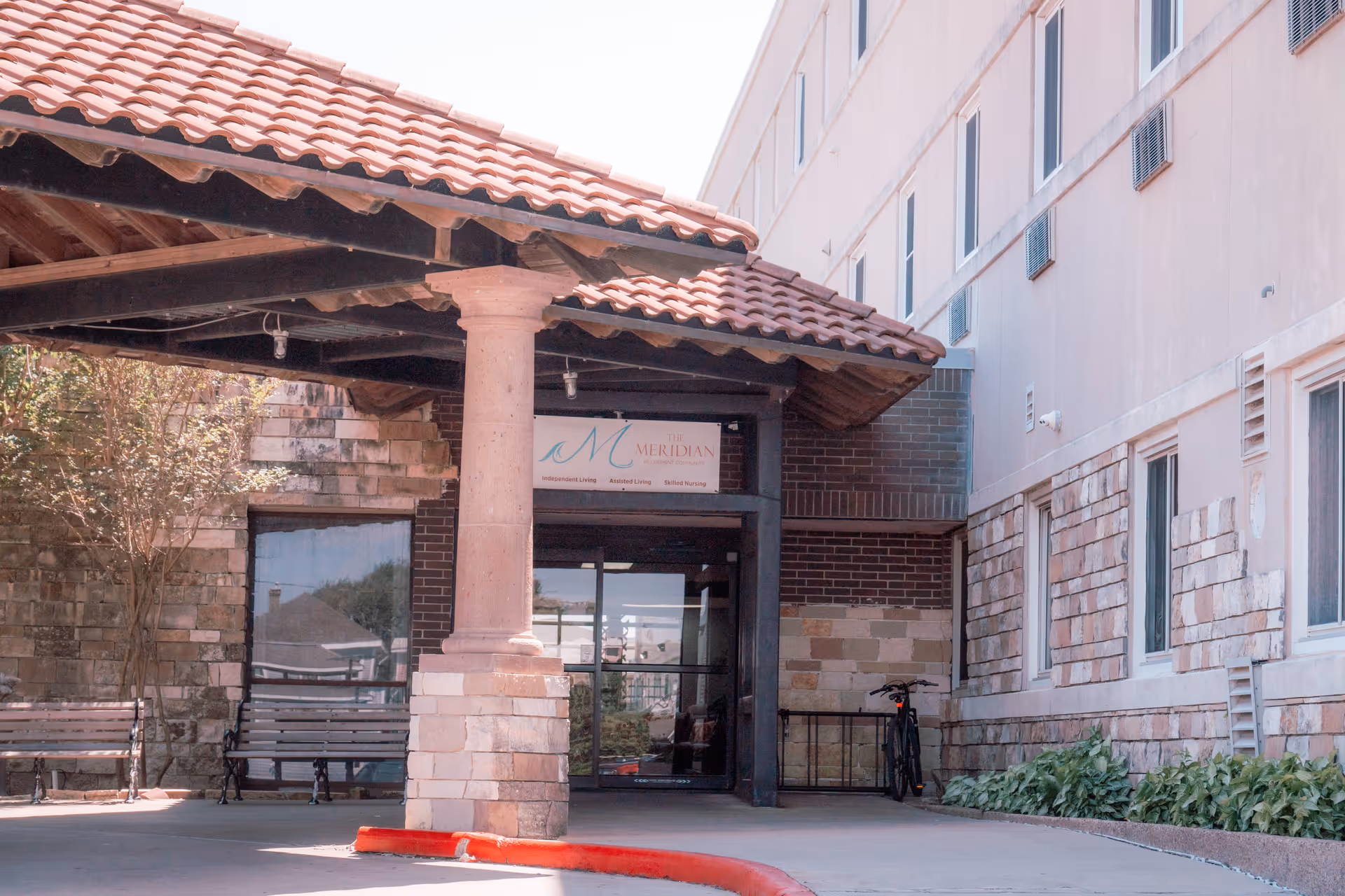 Covered tiled-roof entrance with a stone column in front of The Meridian building, benches and a bicycle by the doorway.