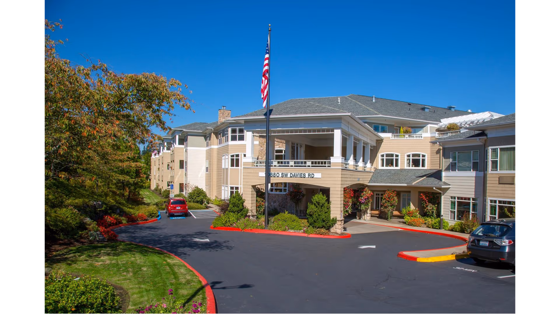 Front entrance of the Hearthstone At Murrayhill senior living building with a flagpole, driveway, parked cars, and landscaped grounds under a clear blue sky.
