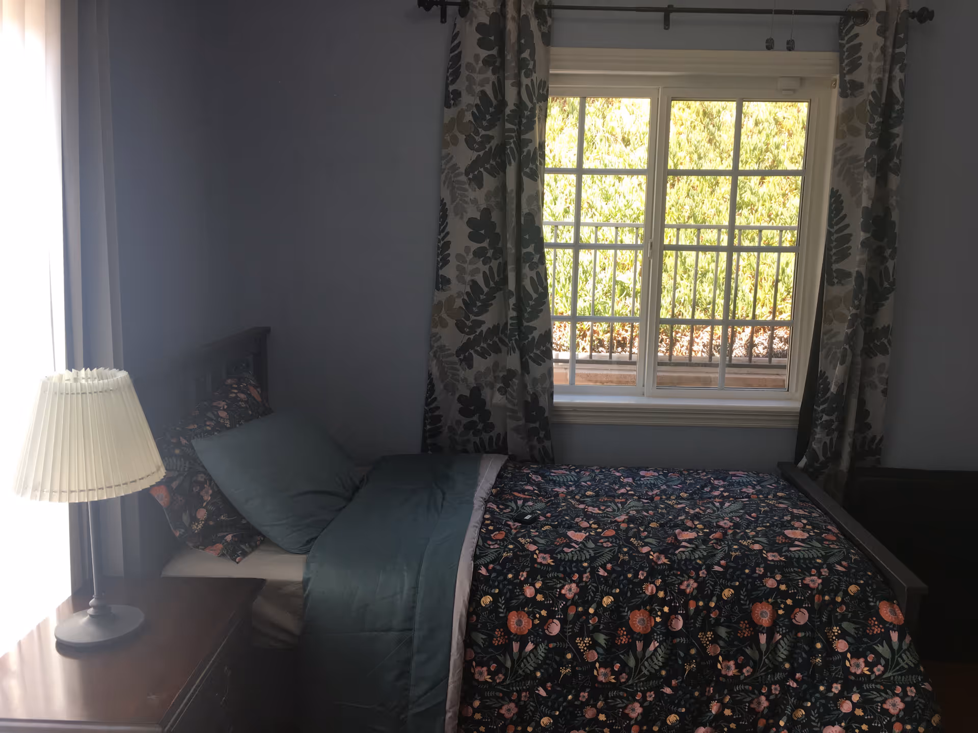 A bedroom with a single bed covered in a floral patterned comforter and green bedding. There is a wooden nightstand next to the bed with a lamp on it. A window with floral curtains lets in natural light and shows greenery outside.