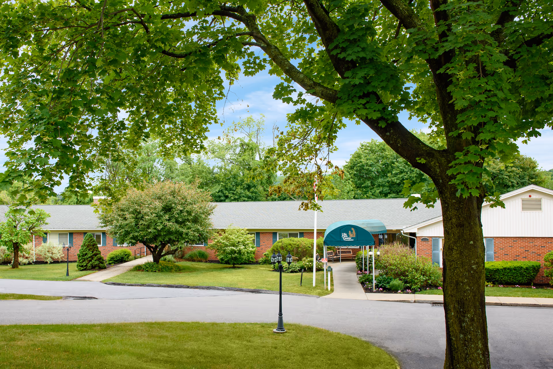 Front exterior of a single-story brick senior living building with a covered entrance, landscaped lawn, and large trees in the foreground.