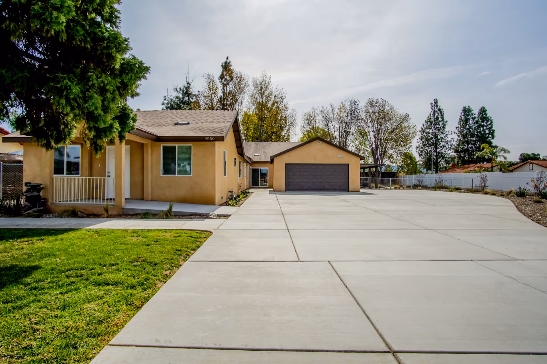 Exterior view of a single-story beige building with a large concrete driveway, a two-car garage, and a small porch with a white railing. There is a green lawn and a large tree on the left side, with other trees and a white fence in the background under a cloudy sky.
