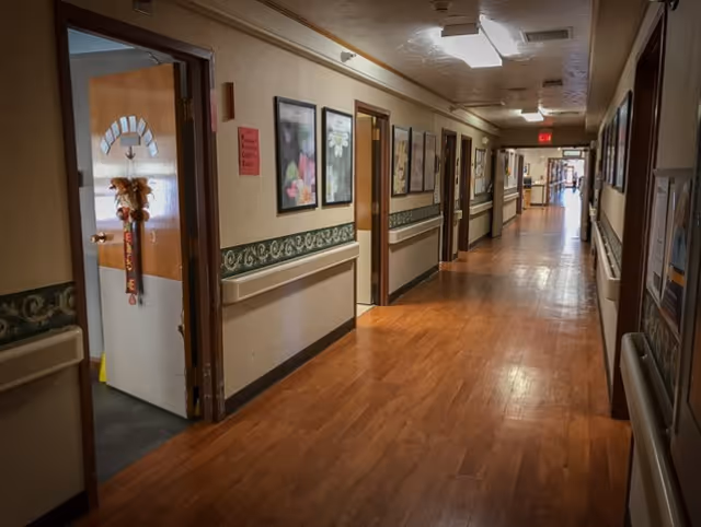 A long hallway in a senior living facility with wooden flooring and beige walls. Several doors line the hallway, some open, and framed floral artwork is hung on the walls. Handrails run along both sides of the hallway. The hallway is well-lit with ceiling lights and an exit sign is visible at the far end.