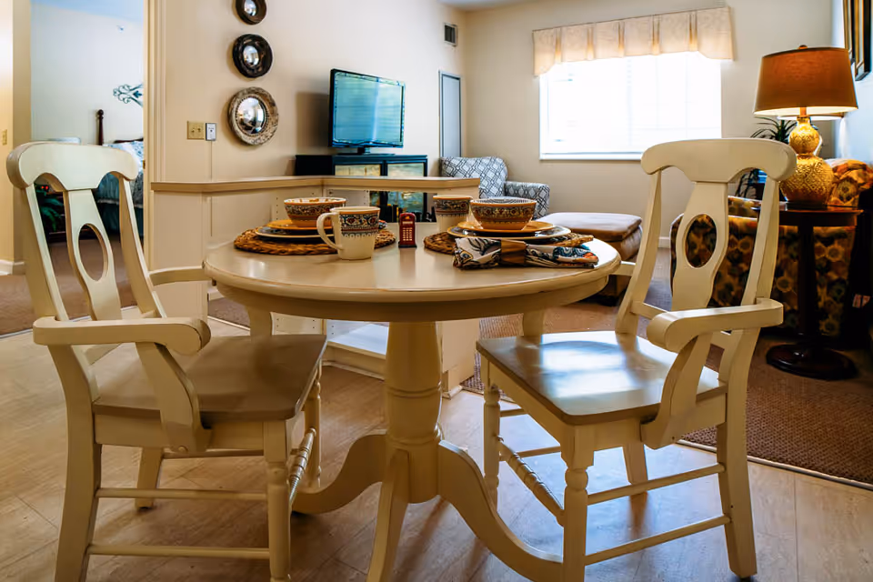 A cozy dining area with a round wooden table set for two with bowls, plates, mugs, and napkins. Two wooden chairs with armrests are positioned at the table. In the background, a living room area is visible with a TV on a stand, an armchair, an ottoman, a side table with a lamp, and a window with blinds and a valance.