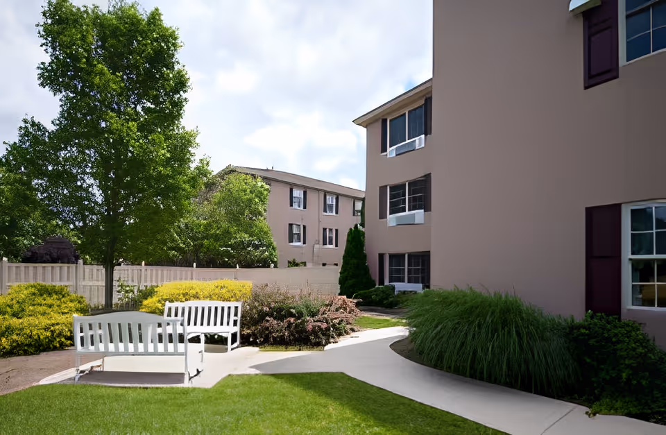 Courtyard with white benches, landscaped shrubs and trees beside the exterior of a multi-story residential building.