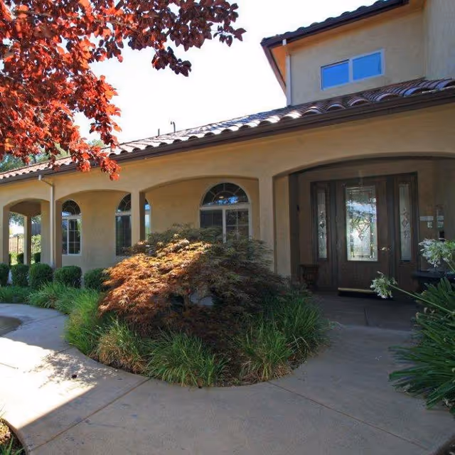 Exterior view of the entrance to a building with beige stucco walls, arched windows, a tiled roof, and a glass front door. There are landscaped bushes and plants along the walkway leading to the entrance, with a tree featuring red leaves partially visible on the left side.
