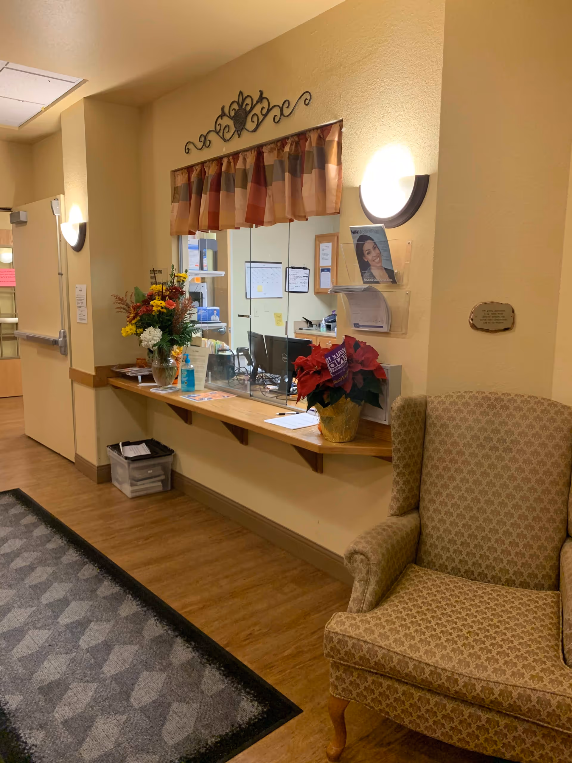 Reception area with a wooden counter and a glass partition. There are two flower arrangements on the counter, one with yellow and red flowers and the other with red poinsettias. Behind the glass, there are computer monitors and office supplies. A patterned armchair is positioned to the right of the counter. The walls are painted beige and decorated with a small curtain and a decorative metal piece above the glass window. The floor has wood-like flooring with a patterned rug near the entrance door.