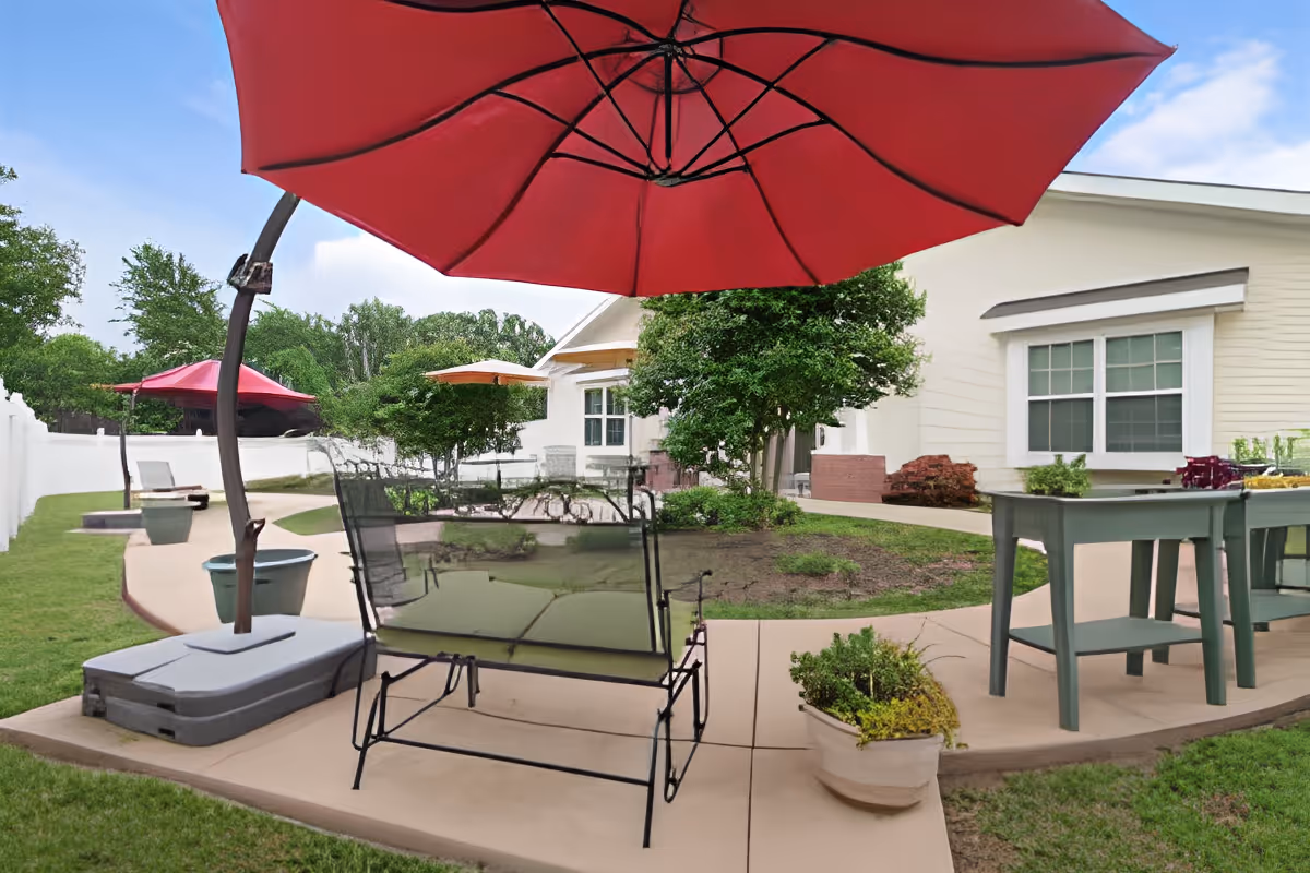 Outdoor patio area with a large red umbrella, metal bench, green table with chairs, potted plants, and a white building with windows in the background under a partly cloudy sky.