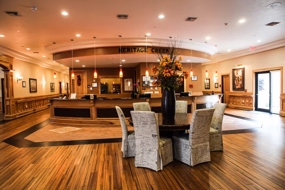 Spacious assisted-living lobby with a round table and large floral centerpiece in front of a curved reception desk under a 'Heritage Creek' sign.