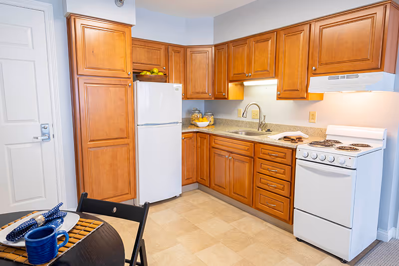 A kitchen area with wooden cabinets, a white refrigerator, a white stove with an overhead vent, a sink with a faucet, and a small dining table set with a blue mug, plate, and napkin. The floor is tiled and the walls are light-colored.