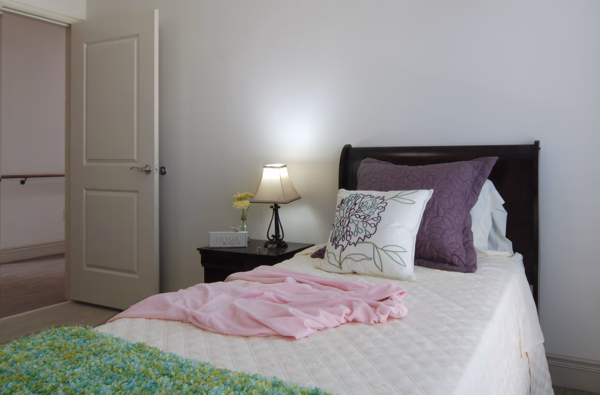 A neatly made single bed with purple pillows, a floral cushion, and a bedside table with a lamp in a bright bedroom.
