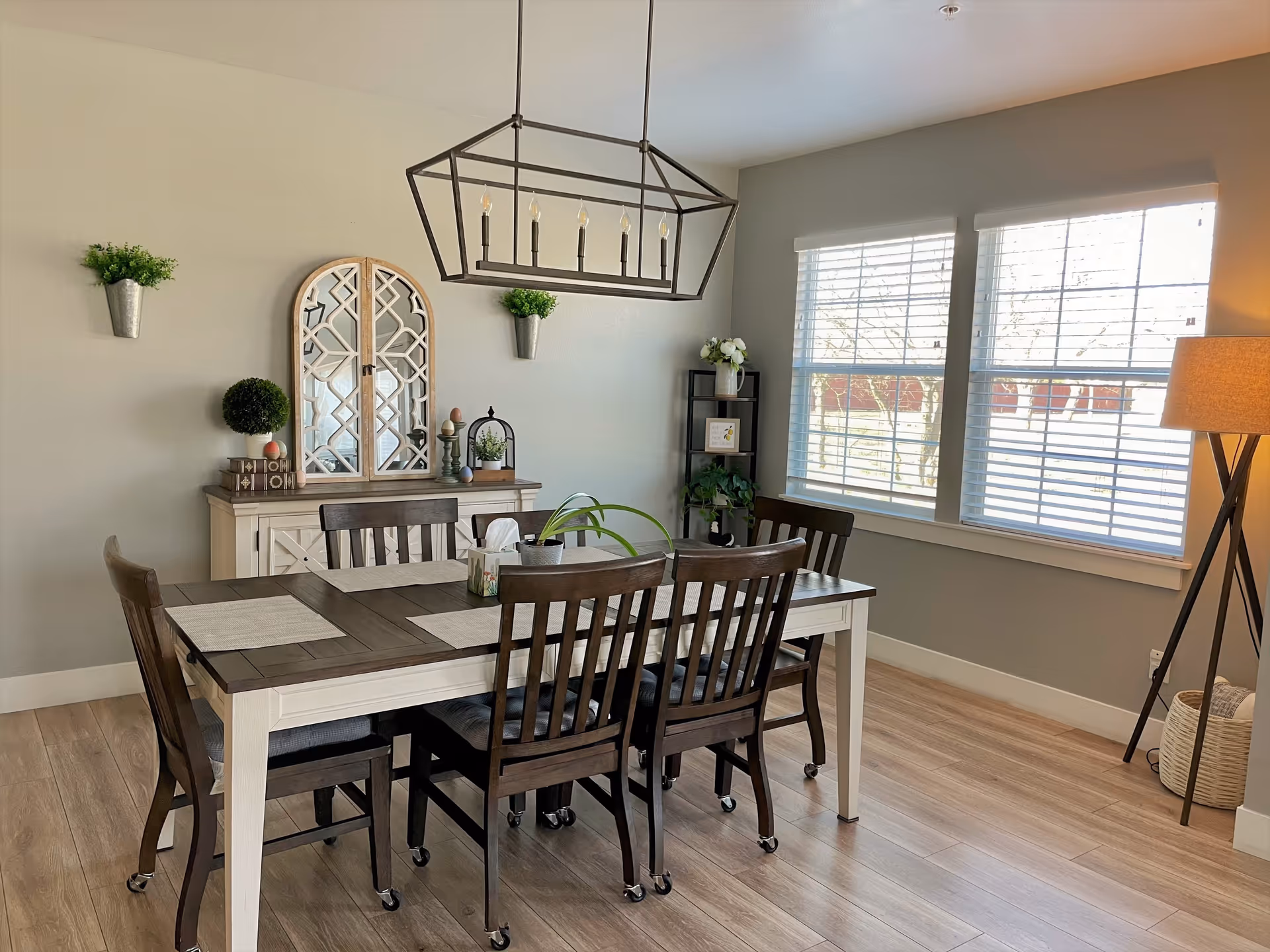 A bright dining room with a wooden table and six chairs on wheels. The table has beige placemats and a small plant centerpiece. On the wall behind the table is a decorative mirror with a wooden frame, flanked by two small potted plants. A modern rectangular chandelier hangs above the table. To the right, there are two large windows with white blinds, and a tall floor lamp with a beige shade stands in the corner. The floor is light wood, and there is a small shelving unit with plants and decor near the windows.