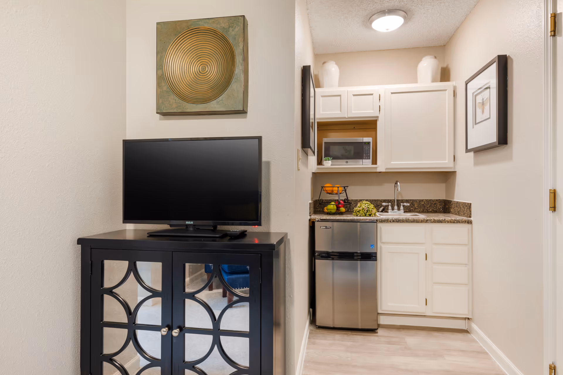 A small studio kitchenette with white cabinets, a mini-fridge, sink and microwave next to a TV on a black cabinet.