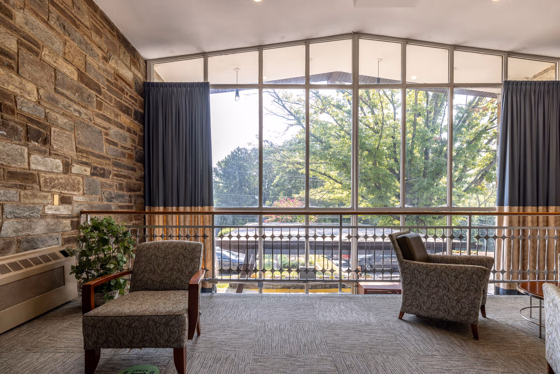 A cozy sitting area with two patterned armchairs facing a large window that offers a view of trees and parked cars outside. The room features a stone wall on the left, a potted plant, and blue curtains framing the window. The floor is carpeted and there is a metal railing in front of the window.