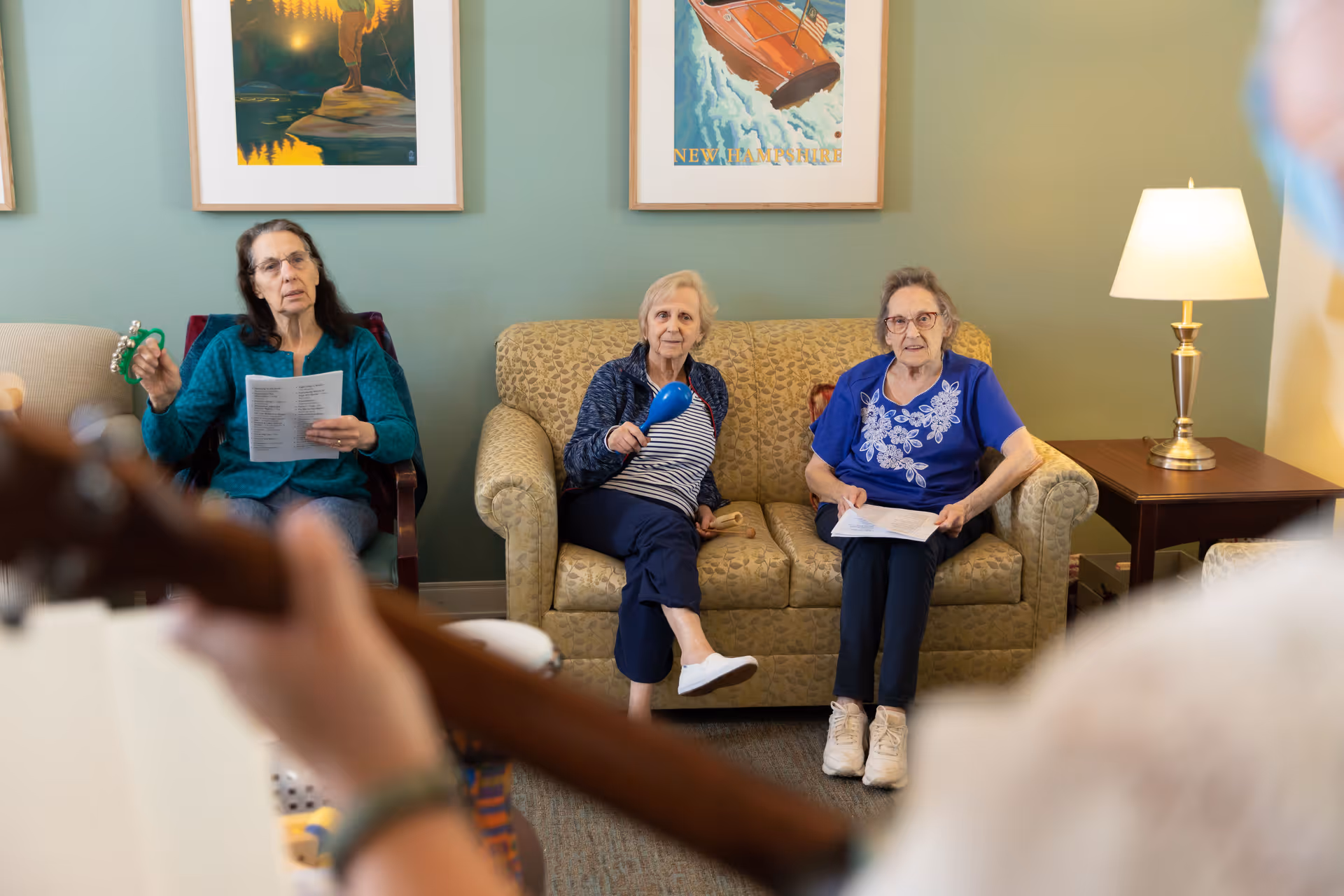 Three elderly women sitting in a living room area, two on a patterned couch and one on a chair, holding musical instruments and papers, while a person in the foreground plays a guitar. The room has green walls with framed artwork and a table lamp on a side table.