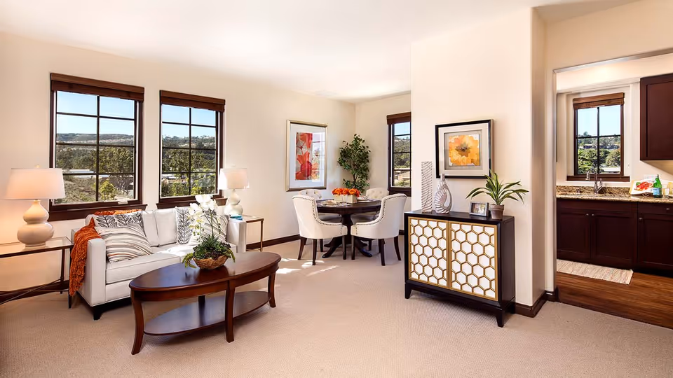 Bright and spacious living area with a white sofa adorned with patterned cushions and an orange throw, a wooden coffee table with a plant centerpiece, two table lamps on side tables, a small dining area with four upholstered chairs around a round table, decorative artwork on the walls, a cabinet with a honeycomb pattern, and a view into a kitchen with dark wood cabinets and granite countertops through an open doorway.