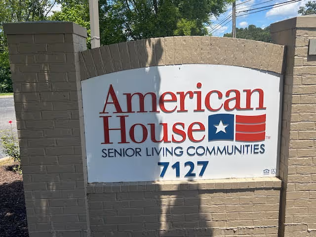 Outdoor sign for American House Senior Living Communities with the address number 7127, mounted on a beige brick structure with trees and a road in the background.