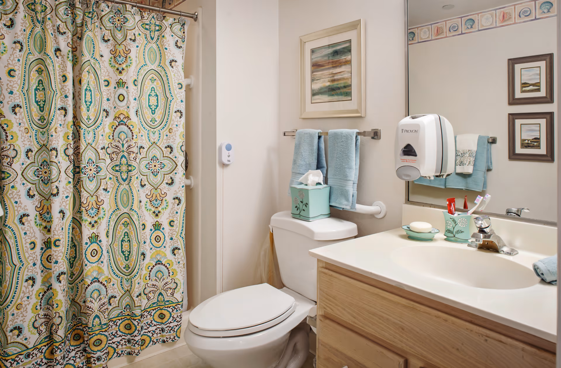 A clean bathroom featuring a toilet with a tissue box on top, a sink with a soap dish, toothbrush holder, and faucet, and a shower with a decorative curtain in green, blue, and beige patterns. Two blue towels hang on a rack above the toilet, and a large mirror reflects part of the room. The walls have a seashell-themed border near the ceiling, and there are framed pictures on the wall.