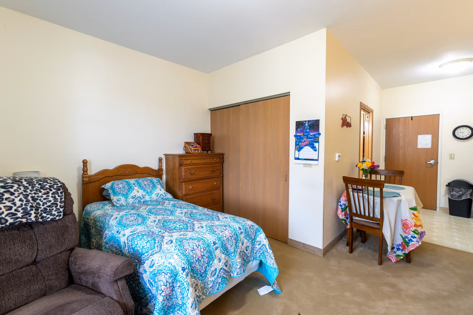 A senior living facility room with a single bed covered in a blue patterned bedspread, a wooden dresser, a brown recliner chair with a leopard print blanket, a small dining table with two chairs and a floral tablecloth, and a door leading to another area. The walls are light-colored and there is a clock and trash can near the door.
