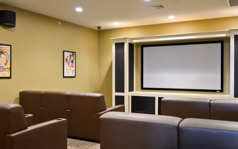 A small theater room with brown leather recliner seats arranged in rows facing a large blank projection screen mounted on a beige wall. The room has yellowish walls with two framed movie posters and recessed ceiling lights.
