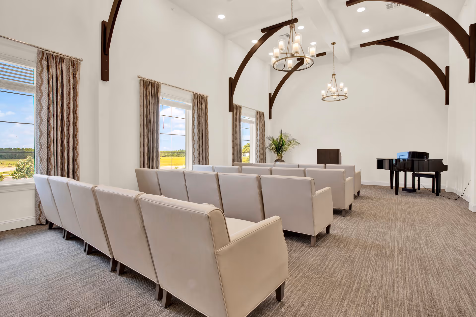 A bright room with beige upholstered chairs arranged in rows facing a black grand piano and a wooden podium. The room has high white walls with dark wooden beams and two chandeliers hanging from the ceiling. Large windows with patterned curtains allow natural light to enter, and a potted plant is placed near the podium.