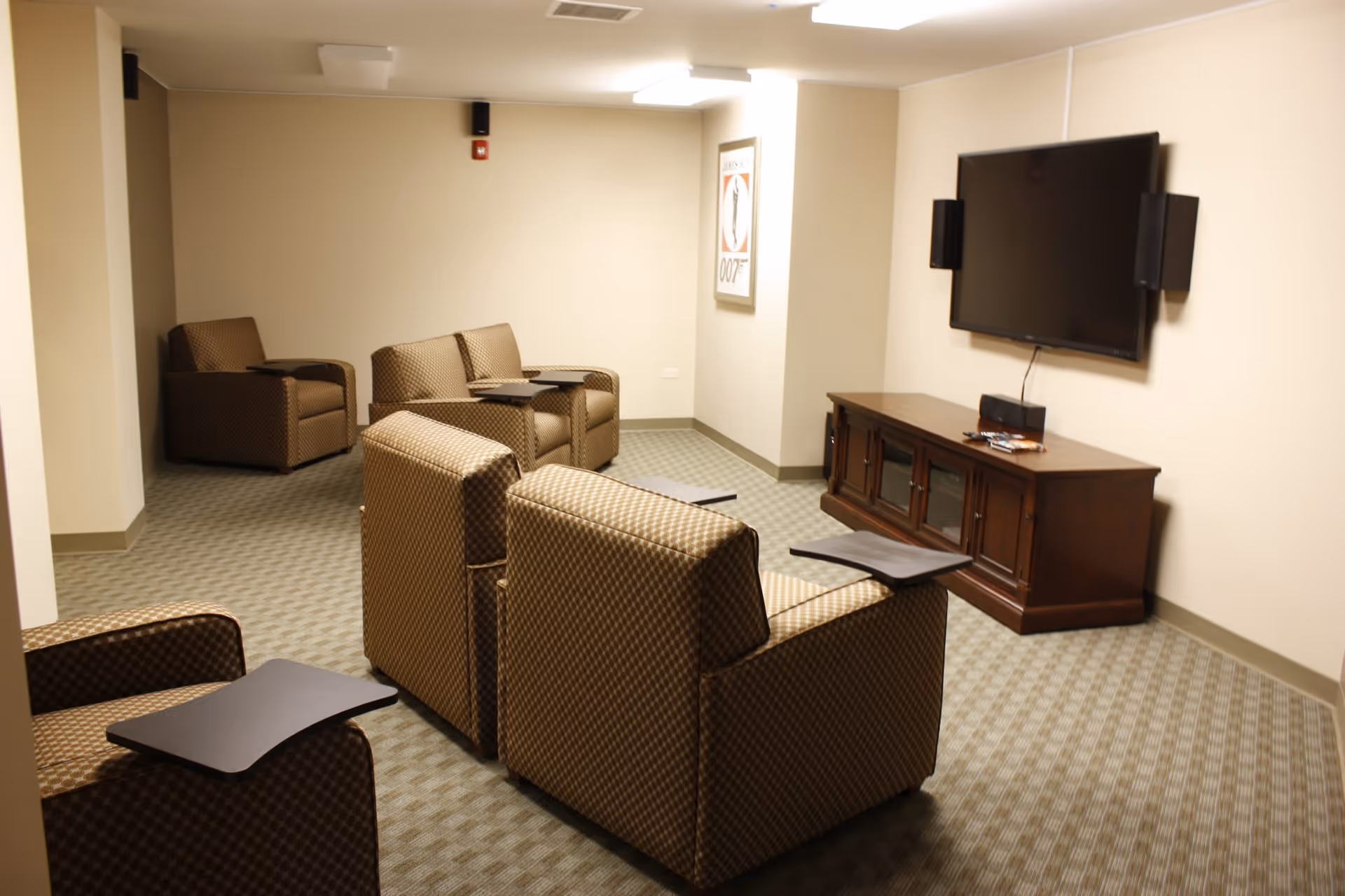 A cozy lounge area with patterned brown armchairs and loveseats arranged facing a wall-mounted flat-screen TV. The room has beige walls, carpeted floor, and a wooden TV cabinet beneath the television. There is a framed poster on the wall to the right.
